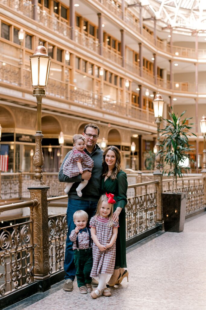 A Cleveland Arcade Family Session by Brittany Serowski Photography. A family of five standing in front of the golden railing and architecture of the Cleveland Arcade in holiday hues of red, green and gray. All are standing close to one another and smiling at the camera.