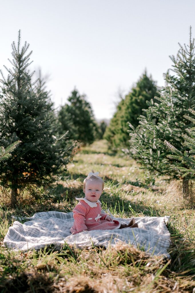 A Cleveland Tree Farm milestone portrait session by Brittany Serowski Photography. A one year old girl in a pink dress and white bow in her hair, sitting on a blanket in front of rows of Christmas Trees, smiling at the camera.