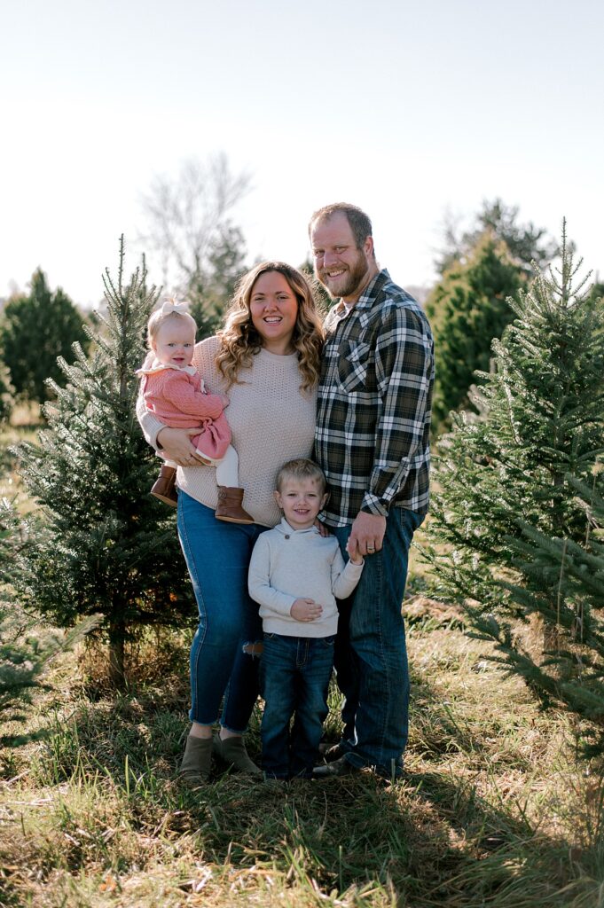 A Cleveland Tree Farm milestone portrait session by Brittany Serowski Photography. A family of four standing in row of trees at a local Christmas Tree Farm, snuggled close to one another smiling at the camera.