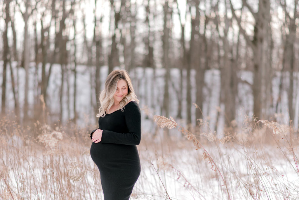An expecting mom wearing a black dress in the snow holding onto her belly as she looks away by Cleveland Maternity Photographer, Brittany Serowski Photography.