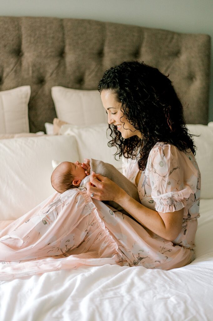 A mother holding her newborn son on her lap in her bedroom with white bedding and a pale pink dress. Photographed by Cleveland newborn photographer, Brittany Serowski Photography.