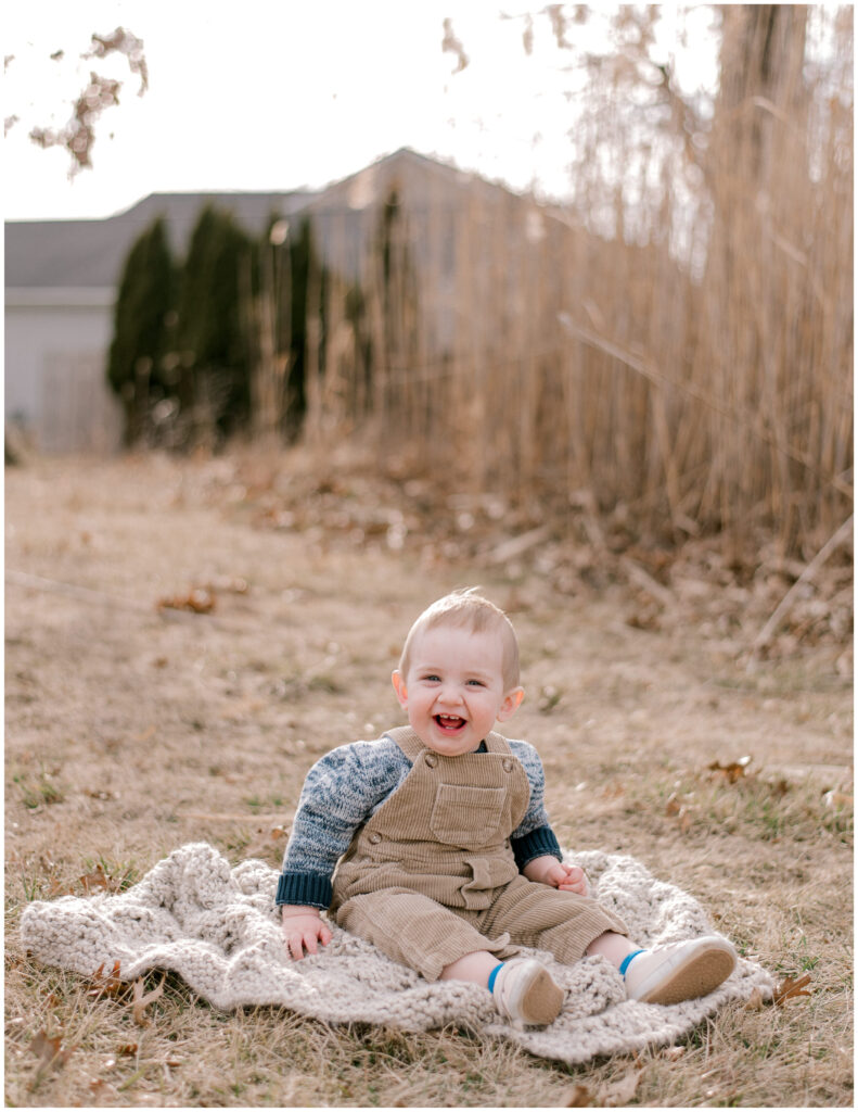 A one year old boy in corduroy overalls and a blue sweater, sitting outside in the winter on a knit blanket smiling at the camera for a one year milestone session by Cleveland Family Photographer, Brittany Serowski Photography.