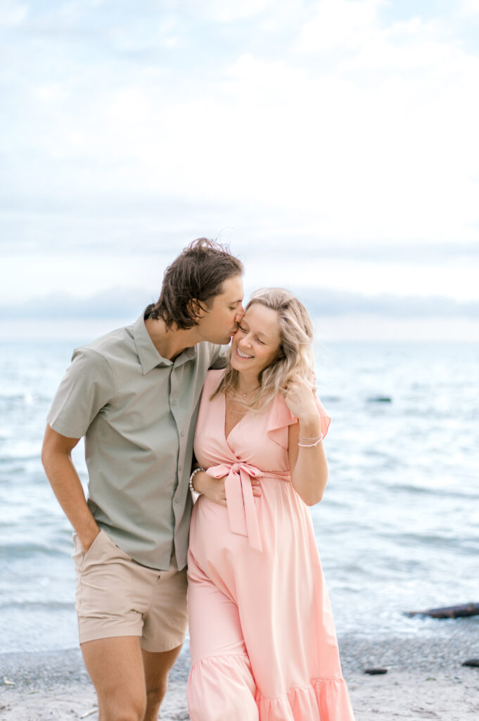 A couple who is expecting, walking arm in arm on the beach with the husband kissing his wife's cheek. Mom is wearing a pink tied dress and dad is in a green button up shirt with khaki pants. Best Time To Book Maternity Portraits by Cleveland Maternity Photographer, Brittany Serowski Photography.