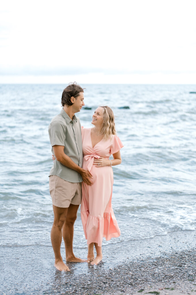 An expecting couple standing on the beach, holding one another close and mom's pregnant belly as they look at one another laughing with the morning beach waves coming in behind them. Best Time To Book Maternity Portraits by Cleveland Maternity Photographer, Brittany Serowski Photography.