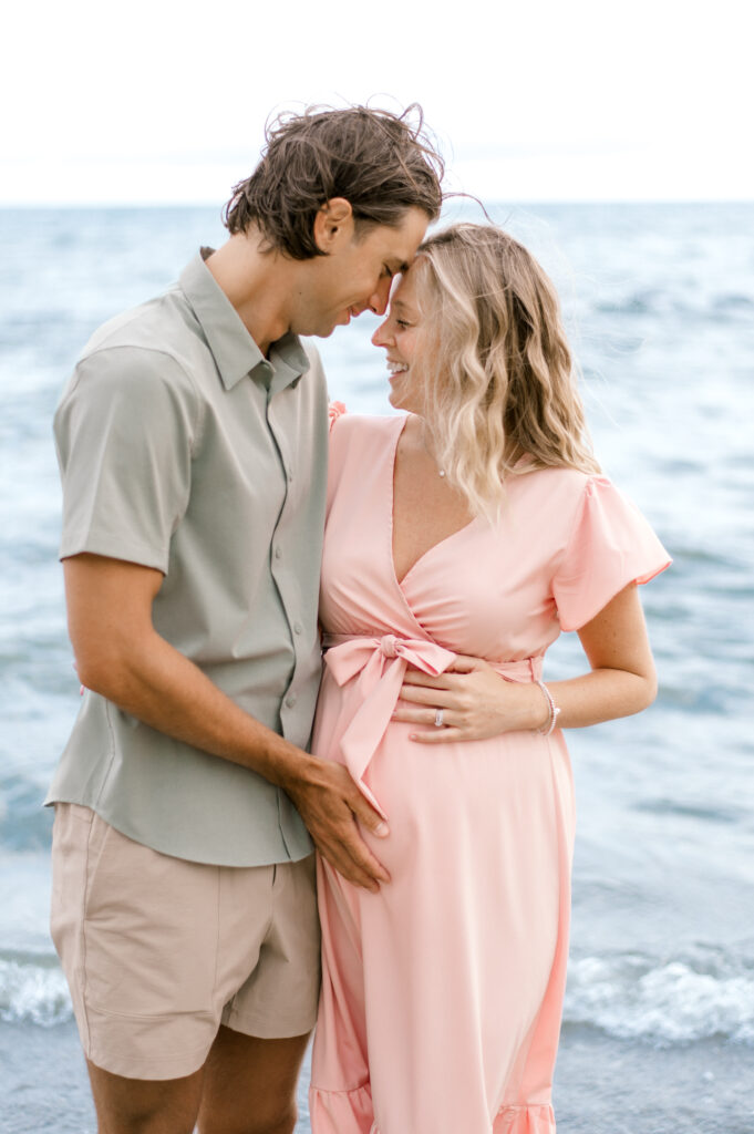 A husband and wife, foreheads together at the lake while smiling and laughing as they hold onto the wife's pregnant belly. Best Time to Book Maternity Photos by Cleveland Maternity Photographer, Brittany Serowski Photography.