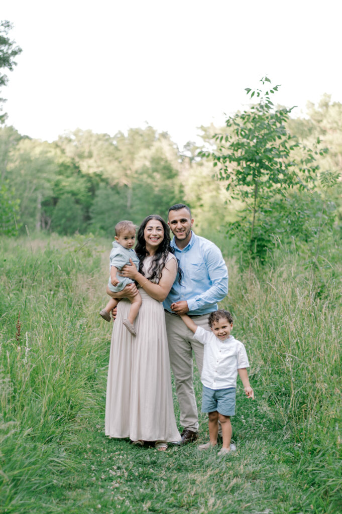 A family of four in cream and blue outfits standing in an open field smiling and looking at the camera while huddled nice and close together by Brittany Serowski Photography for her Fall & Holiday Cleveland Minis.