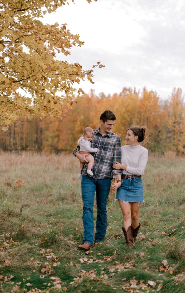 A young family of three in a fall setting of an open field with yellow and orange leaves on the trees behind them as they smile at one another while walking hand in hand, with dad holding their baby girl on his hip. Couple and child is in neutral fall colors in part of Brittany Serowski Photography's fall seasonal style guide on what to wear for fall family photos.