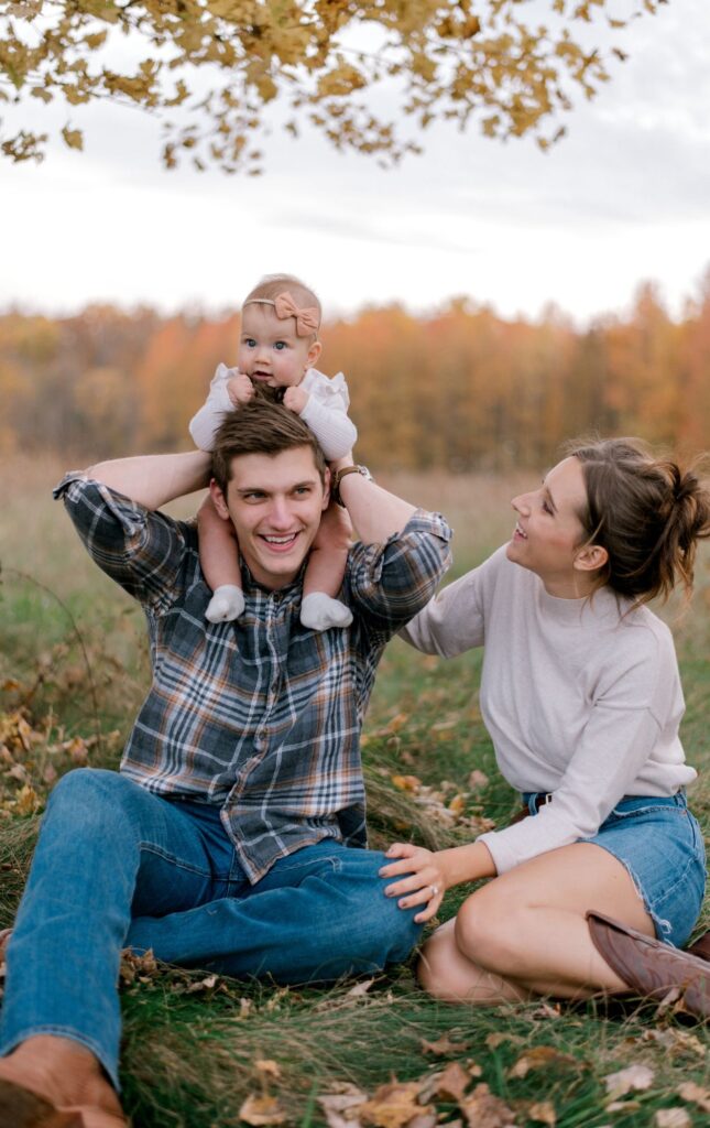 A young family of three in a fall setting of an open field with yellow and orange leaves on the trees behind them as they smile at each other while sitting on the ground, with dad holding their baby girl on his shoulders. Couple and child is in neutral fall colors in part of Brittany Serowski Photography's fall seasonal style guide on what to wear for fall family photos.