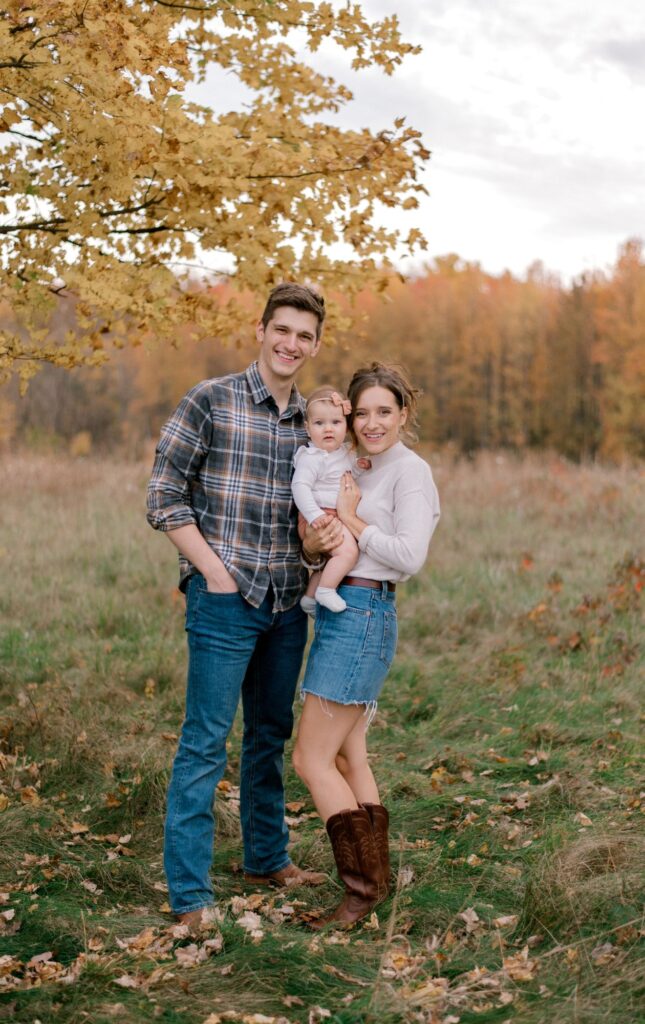 A young family of three in a fall setting of an open field with yellow and orange leaves on the trees behind them as they smile at the camera, with mom and dad holding their baby girl in between them. Couple and child is in neutral fall colors in part of Brittany Serowski Photography's fall seasonal style guide on what to wear for fall family photos.