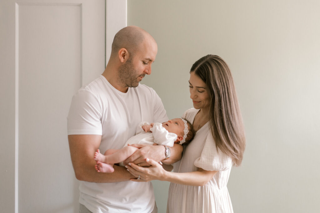 A new father, in a white Tshirt, holding his newborn daughter while gazing at her. Mom is standing at their side while also holding onto their newborn and looking down at her too. Captured by Cleveland Newborn Photographer, Brittany Serowski Photography.