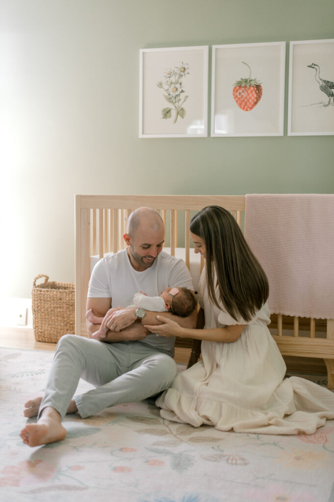 New parents sitting in front of a natural wood crib on the floor, while holding their newborn daughter in her green nursery with Etsy framed prints on the wall behind them. Captured by Cleveland Newborn Photographer, Brittany Serowski Photography.