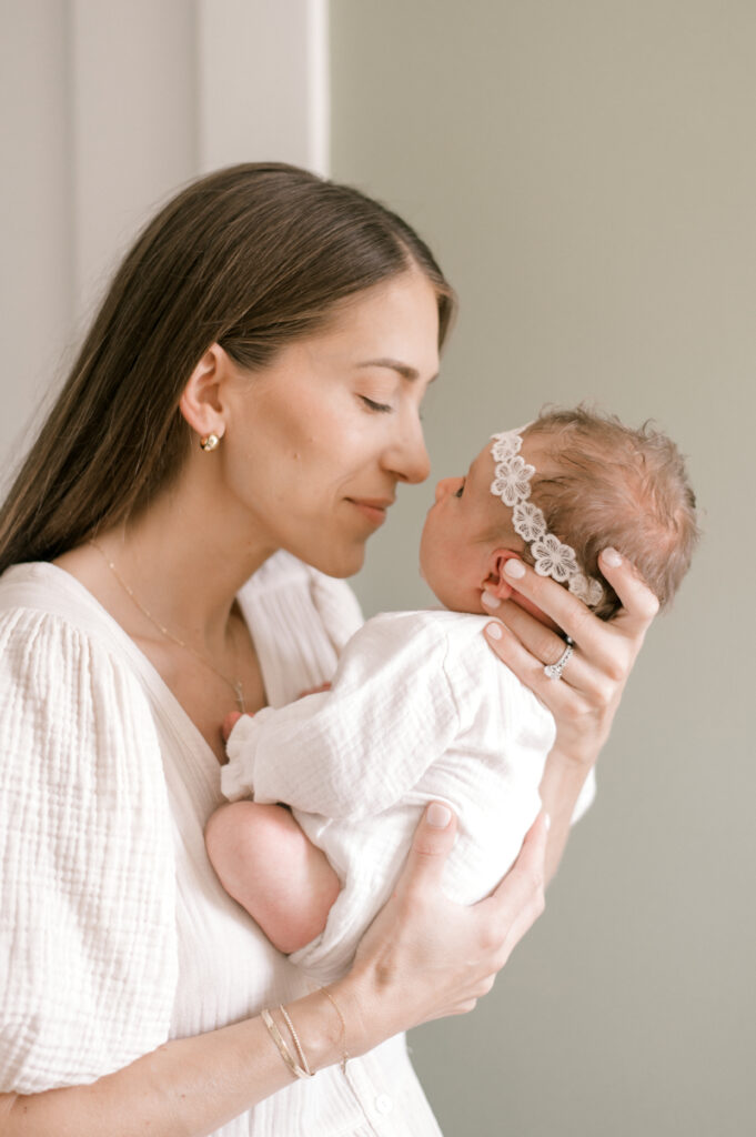 A new mom, in a cream colored dress, nose to nose with her newborn daughter who is wearing a matching cream colored onesie with a floral headband. Captured by Cleveland Newborn Photographer, Brittany Serowski Photography.