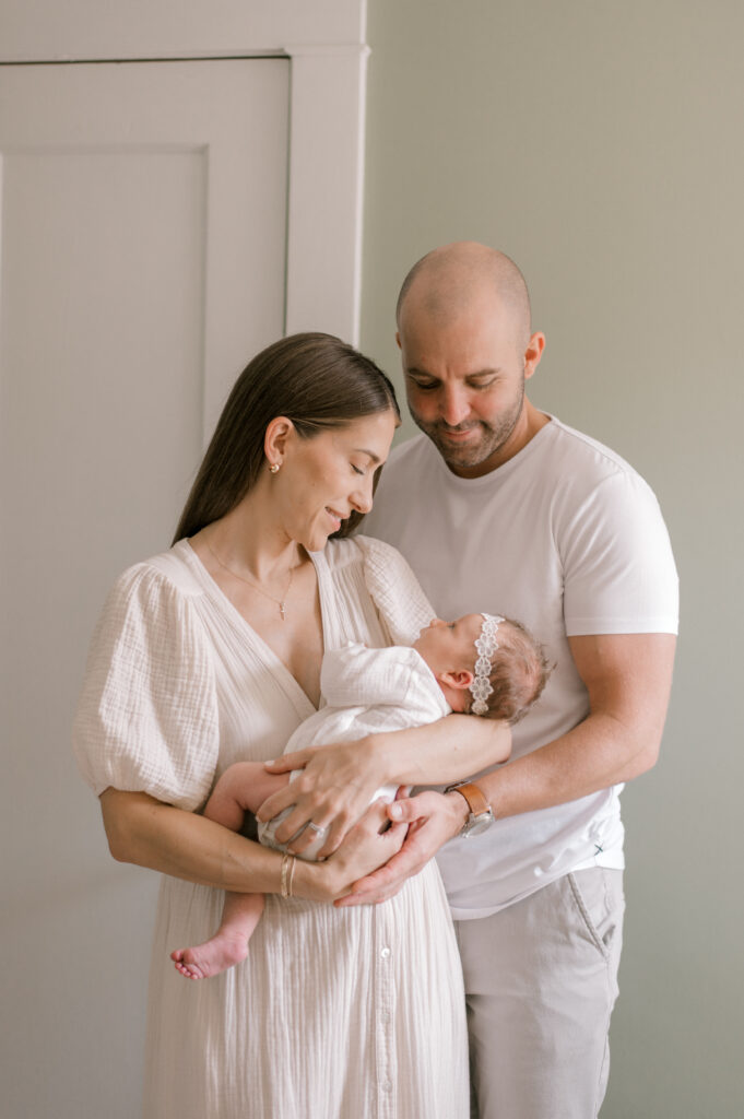 A new mom, in a neutral cream colored dress, holding her newborn daughter who is in a cream long sleeve onesie. While they are gazing at each other, dad standing behind holding them both in his arms, while also looking at his newborn daughter. Captured by Cleveland Newborn Photographer, Brittany Serowski Photography.