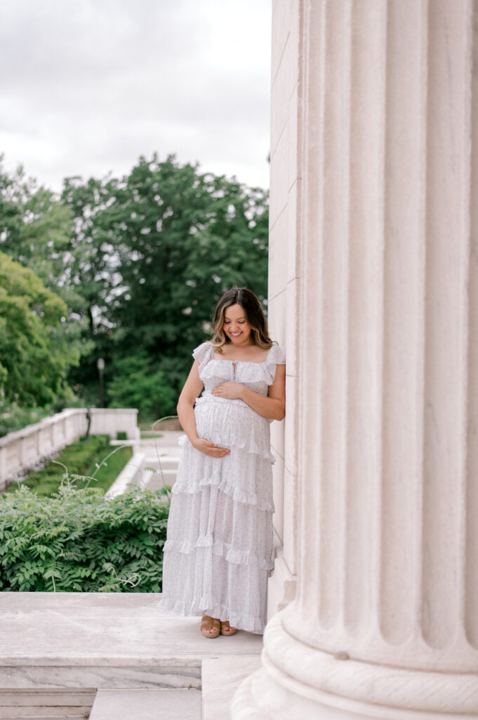 An expecting mom in a white short sleeve and floral full length dress, leans again the pillars of the Cleveland Museum of Art, while holding her belly and looking down at her expecting bump. Captured by Cleveland Maternity Photographer, Brittany Serowski Photography.