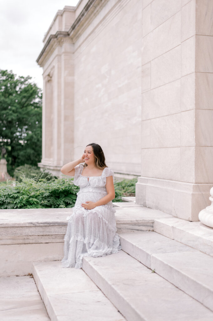 An expecting mom in a white short sleeve and floral full length dress, sits on the steps of the Cleveland Museum of Art, while holding her belly and looking off into the distance. Captured by Cleveland Maternity Photographer, Brittany Serowski Photography.