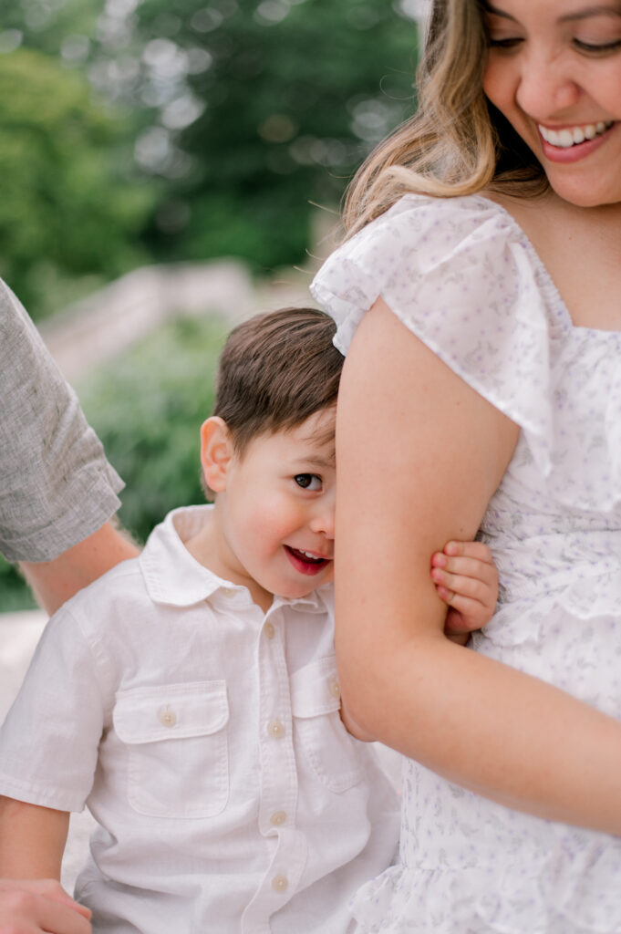 A toddler boy, hiding behind mom's arm and peeking at the camera as they sit on the steps on the Cleveland Museum of Art. Captured by Cleveland Maternity Photographer, Brittany Serowski Photography.