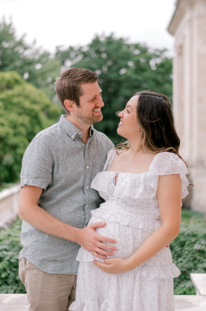 An expecting mom and dad, in neutral, light palette clothing, hold onto her expecting bump together as they look at each other while standing in front of the Cleveland Museum of Art, while holding her belly and looking off into the distance. Captured by Cleveland Maternity Photographer, Brittany Serowski Photography.