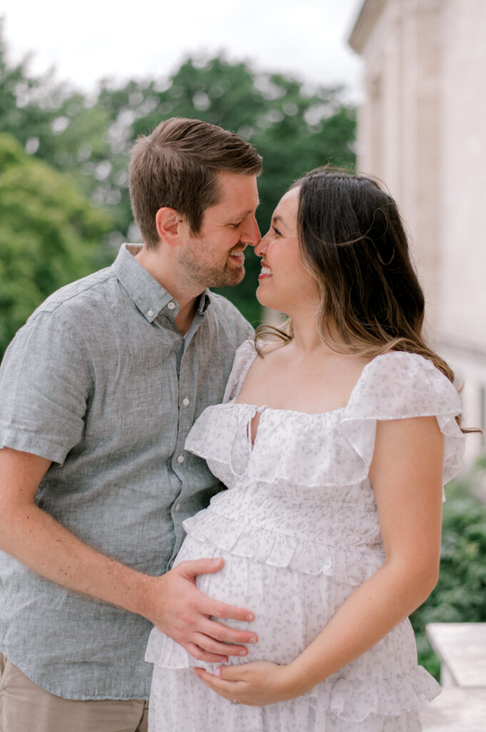 An expecting mom and dad, in neutral, light palette clothing, hold onto her expecting bump together as they look at each other while standing in front of the Cleveland Museum of Art, while holding her belly and looking off into the distance. Captured by Cleveland Maternity Photographer, Brittany Serowski Photography.