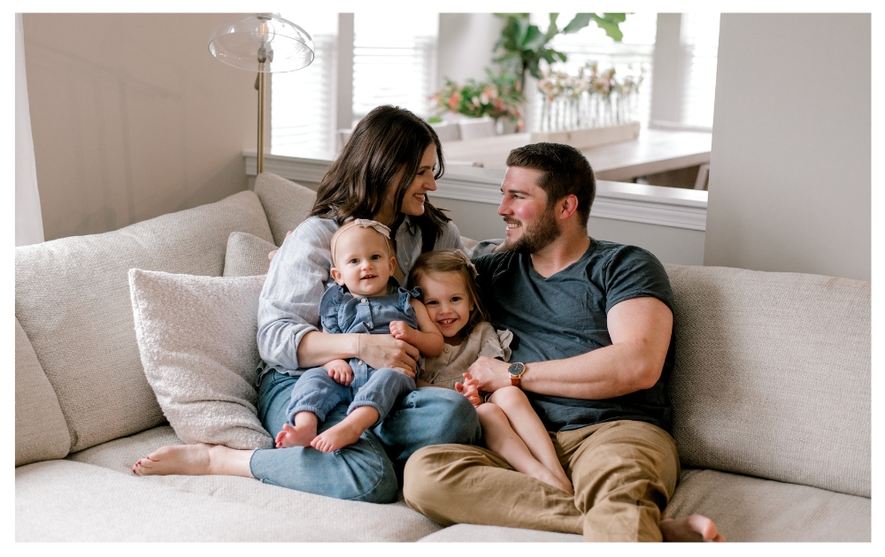 A family of four sitting together on the couch snuggled up, while mom and dad smile at each other and their two young daughters look ahead, smiling at the camera. The family is in neutral blue and cream clothing in their light and airy home. Captured by Cleveland Family Photographer, Brittany Serowski Photography.