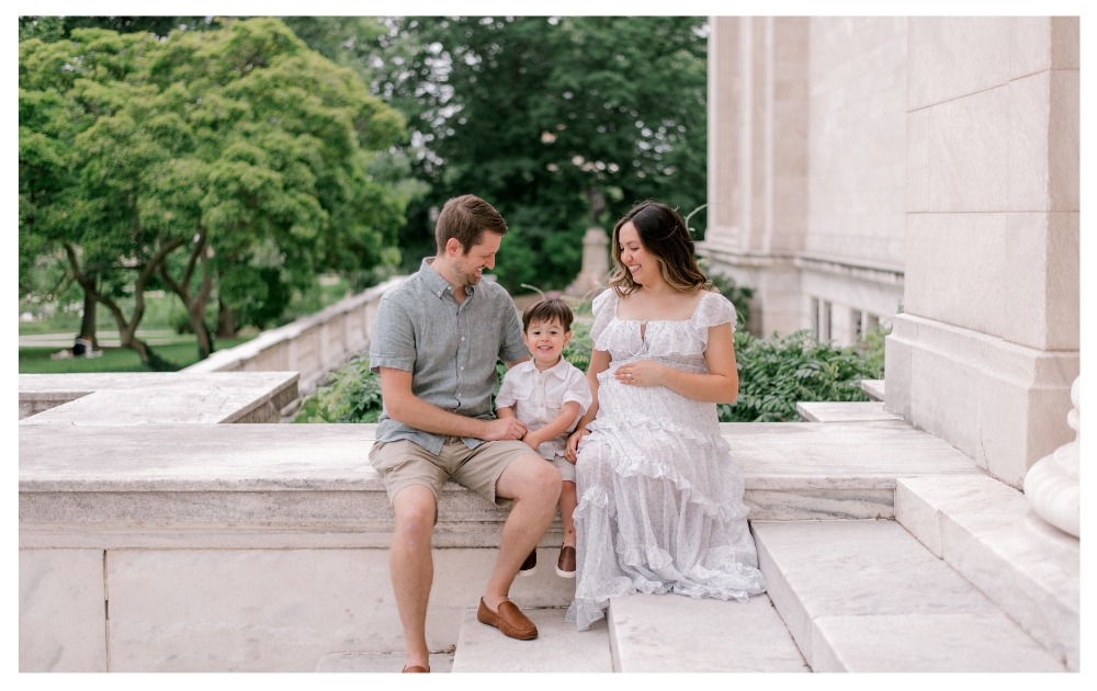 A family of three sitting on the steps on the Cleveland Museum of Art while mom holds her belly as she is expecting their second child. Mom and dad are both looking down at their toddler son, while he looks onward to the camera smiling. Captured by Cleveland Maternity Photographer, Brittany Serowski Photography.