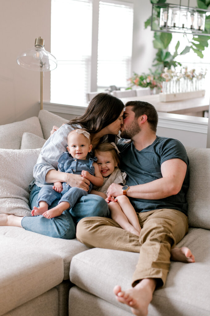 A family of four sitting together on the couch snuggled up, while mom and dad kiss each other and their two young daughters look ahead, smiling at the camera. The family is in neutral blue and cream clothing in their light and airy home. Captured by Cleveland Family Photographer, Brittany Serowski Photography.