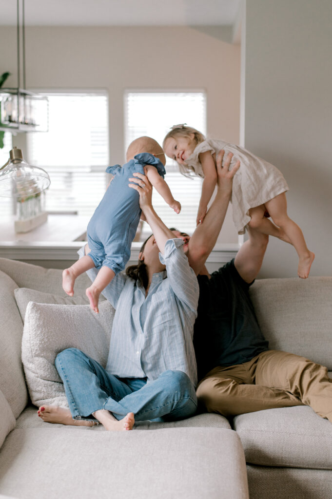 A family of four, sitting together on the couch as mom and dad play airplane with their two young daughters. The family is wearing a blue and white cohesive neutral color palette in their light and airy home. Captured by Cleveland Family Photographer, Brittany Serowski Photography.