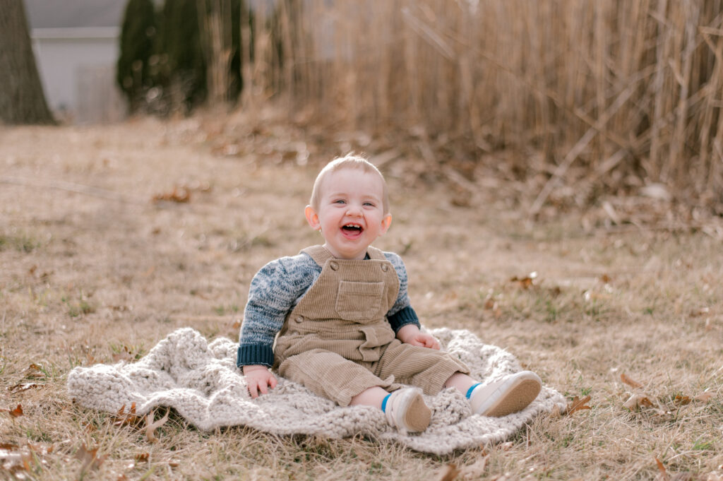 A one year old boy, sitting outside in front of pampas grass with a camel colored corduoroy overalls while smiling and looking right at the camera. Photographed by Cleveland Baby Photographer, Brittany Serowski Photography.