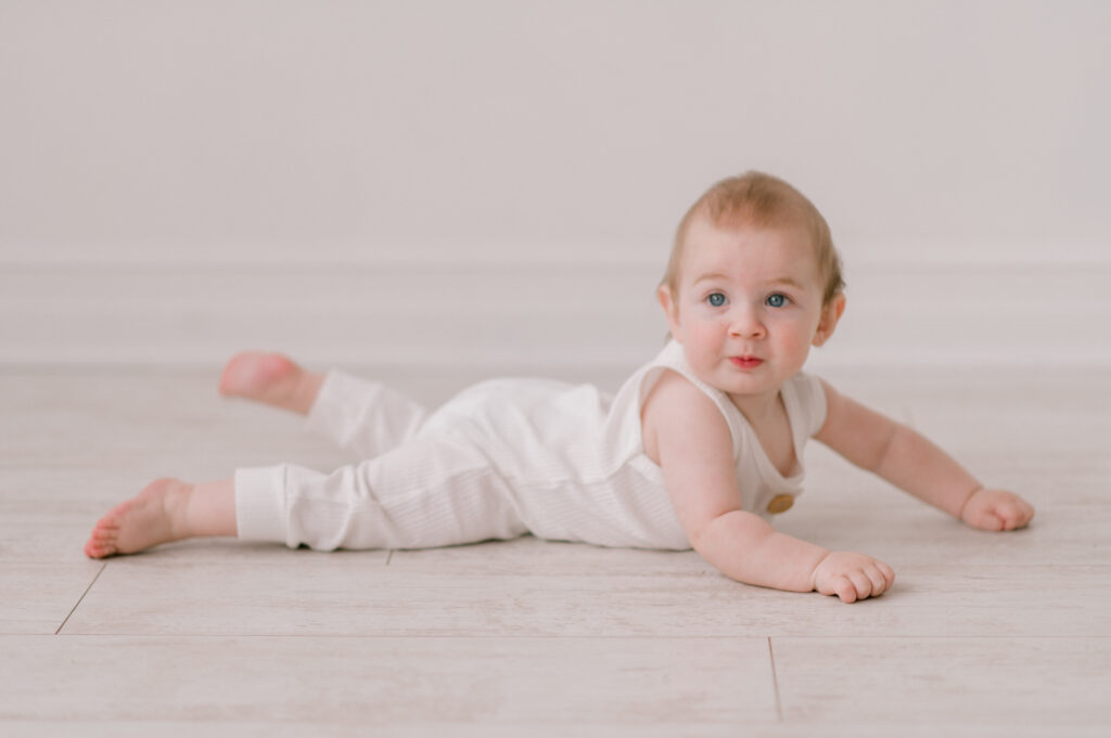 A baby boy in a white romper, laying on his belly in an all white studio in Cleveland, looking at the camera with his arms out in front of him. Photographed by Cleveland Milestone Photographer, Brittany Serowski Photography.