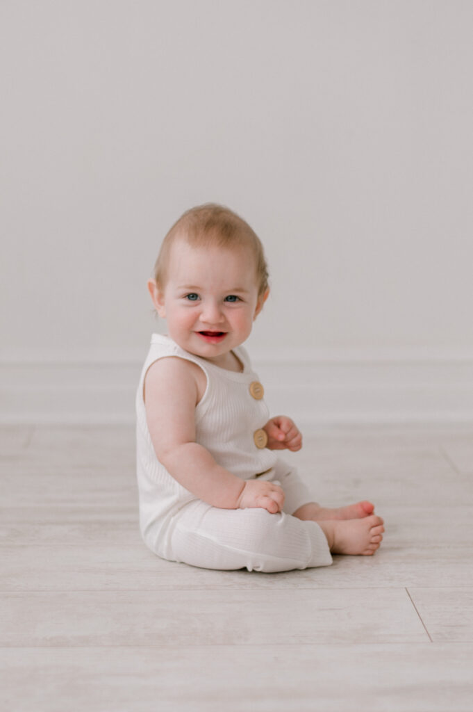 A baby boy in a white romper, sitting up in an all white studio in Cleveland, looking at the camera. Photographed by Cleveland Milestone Photographer, Brittany Serowski Photography.