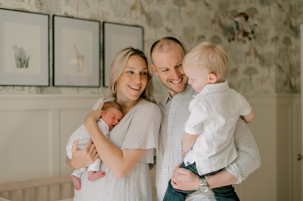 A family of four, standing in a neutral nursery for their baby boy. Mom is holding her newest baby close to her chest, while dad is holding their toddler son in his arms. They are all looking at their oldest son and smiling with delight. Captured by Cleveland Newborn Photographer, Brittany Serowski Photography.