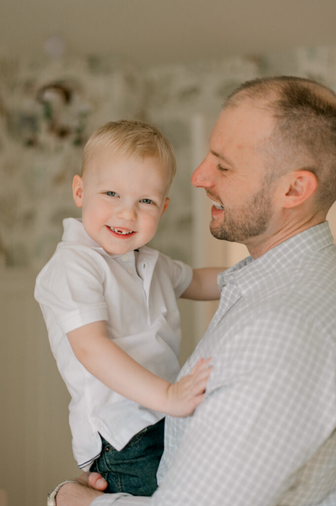 A toddler boy, being held by his dad is smiling at the camera while standing in a neutral nursery. Dad and son are both in neutral shirts. Captured by Cleveland Newborn Photographer, Brittany Serowski Photography.