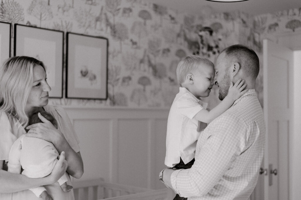 A black and white image of a dad and toddler son holding one another's faces while mom is standing off to the side watching them with joy as she holds her newborn baby close to her chest. Captured by Cleveland Newborn Photographer, Brittany Serowski Photography.