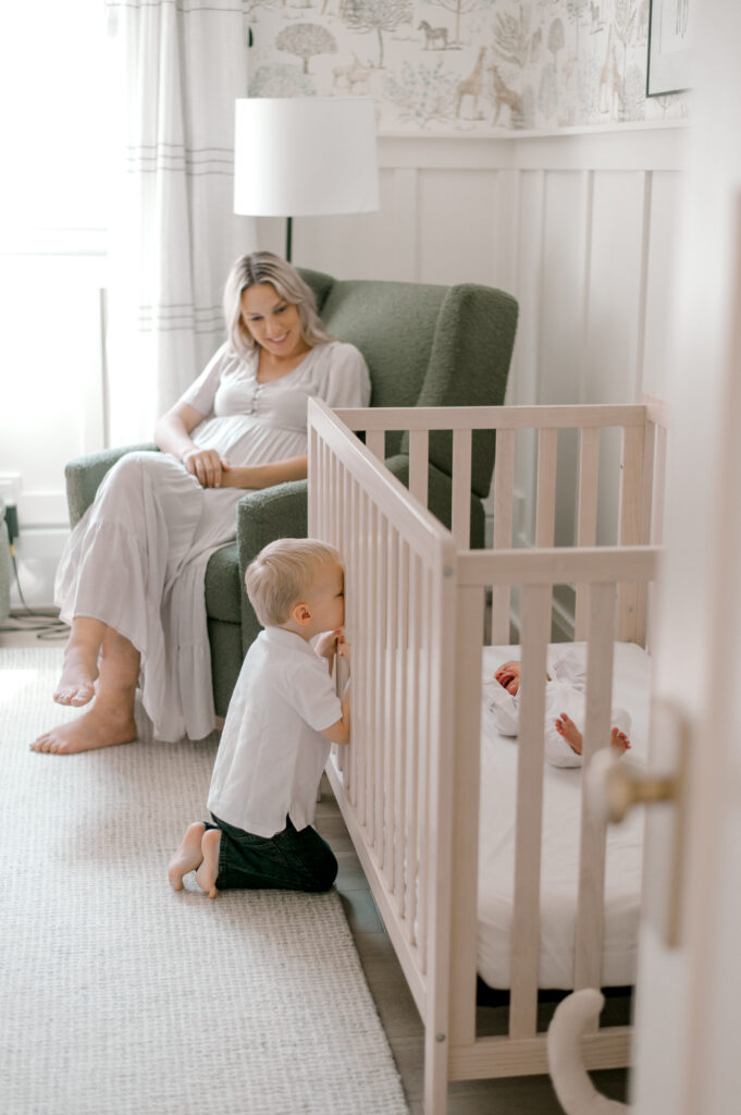 A toddler boy looking through the slats of a crib onto his newborn brother. Mom is sitting in a rocking chair to the back as she watches them both. Captured by Cleveland Newborn Photographer, Brittany Serowski Photography.