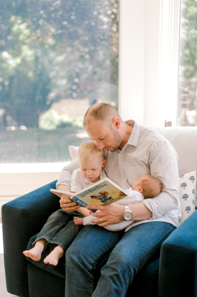 A dad is sitting in a light filled room while holding his two sons, a toddler and a newborn and reading to them Little Blue Truck. Captured by Cleveland Newborn Photographer, Brittany Serowski Photography.