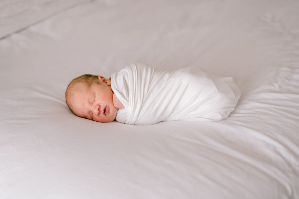 A newborn baby wrapped in a white swaddle on a white bed while sleeping. Photographed by Cleveland Newborn Photographer, Brittany Serowski Photography.