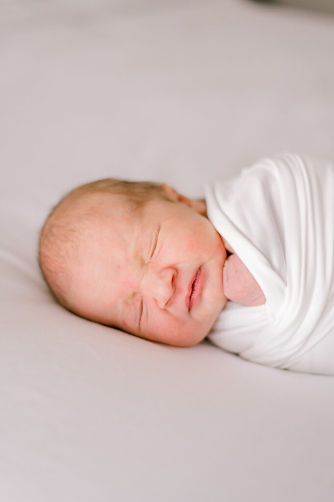 A newborn baby wrapped in a white swaddle on a white bed while sleeping. Photographed by Cleveland Newborn Photographer, Brittany Serowski Photography.