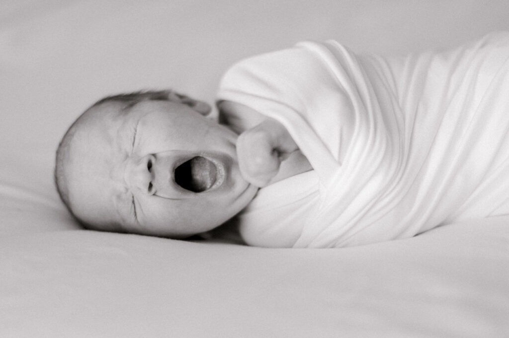 A newborn baby wrapped in a white swaddle on a white bed while yawning. Photographed by Cleveland Newborn Photographer, Brittany Serowski Photography.