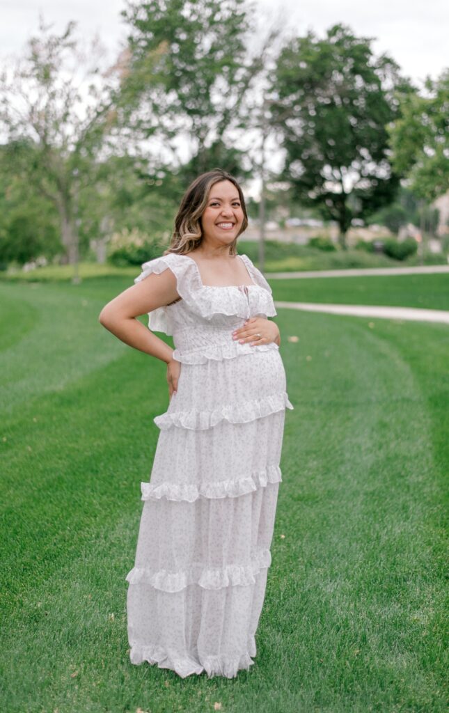 An expecting mother in a white floral full length maternity dress, on the lawn of the Cleveland Museum of Art, smiling at the camera as she holds on to her expecting belly. Captured by Cleveland Maternity Photographer, Brittany Serowski Photography.