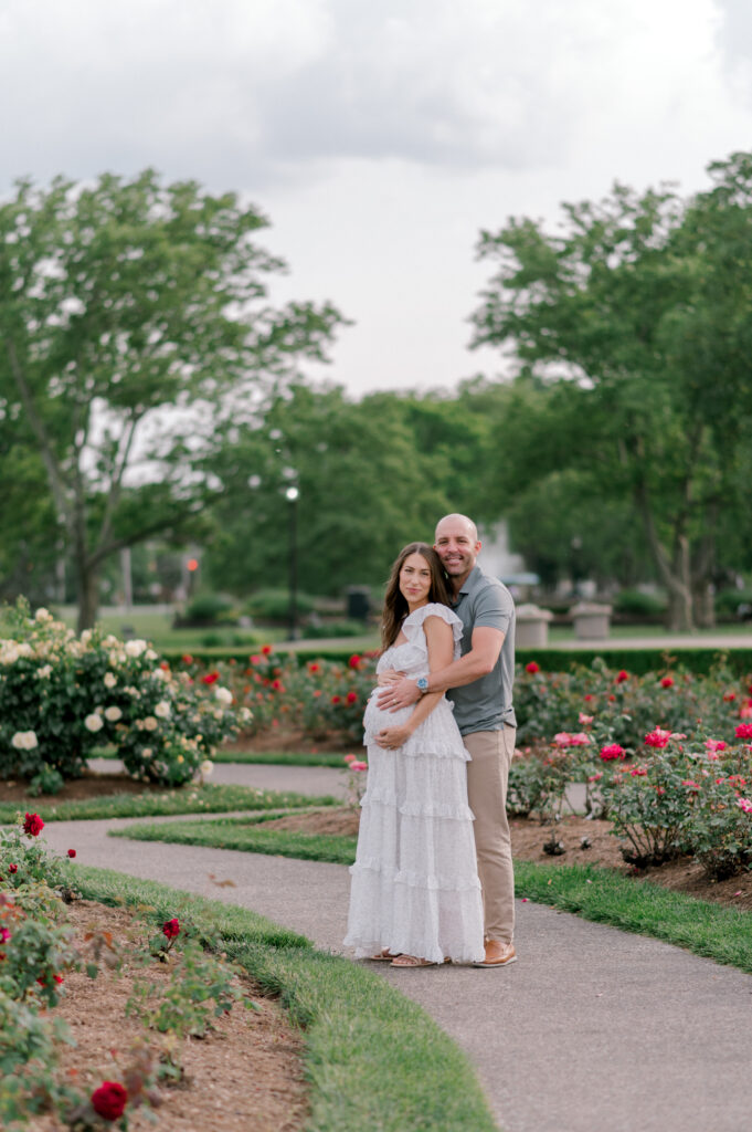 An expecting mom and dad standing in the rose garden at Lakeview Beach in Lorain, OH. Mom is in a white floral floor length dress as dad is in a sage green button up shirt and khaki pants. They are snuggled up to each other as they smile at the camera and hold onto mom's belly bump. Photographed by Cleveland Maternity Photographer, Brittany Serowski Photography.