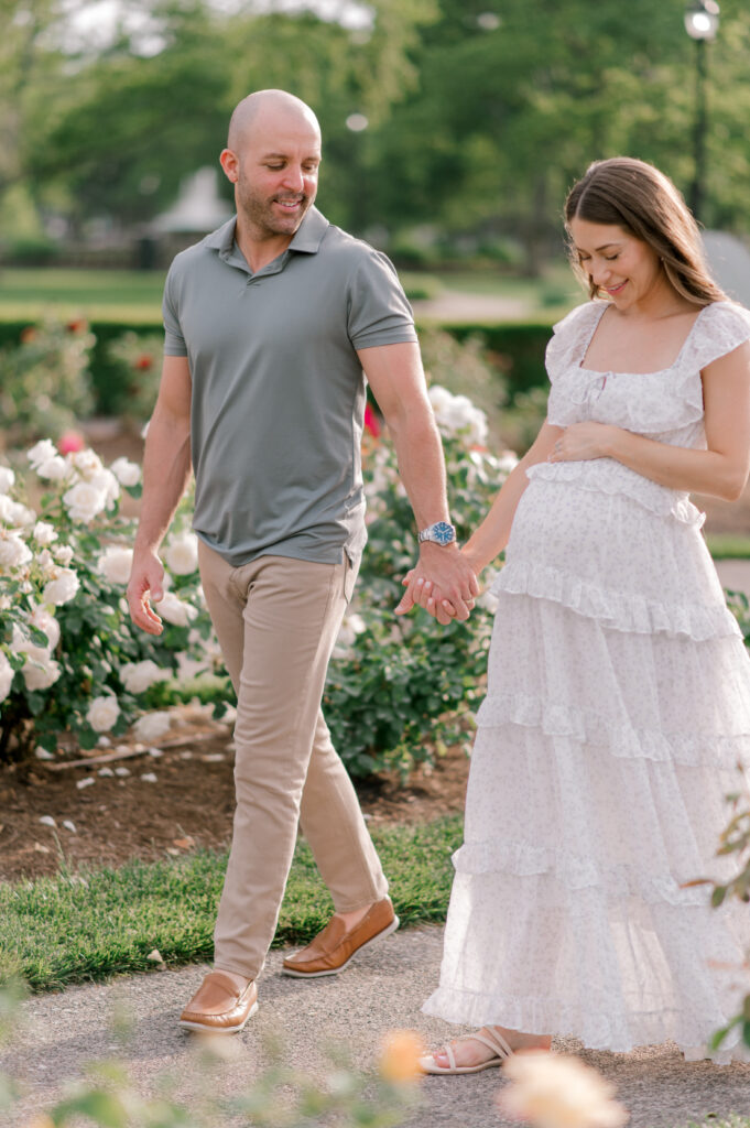An expecting couple, walking hand in hand as they look onto mom's belly bump in the rose garden at Lakeview Beach in Lorain, OH. Photographed by Cleveland Maternity Photographer, Brittany Serowski Photography.