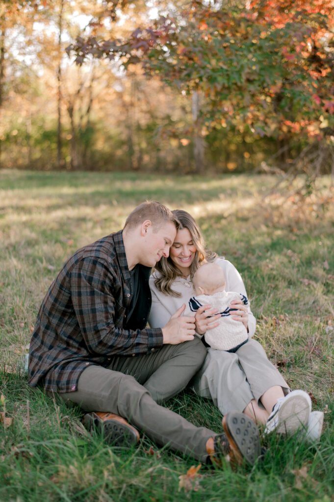 A young family of three, sitting in an open field in the beginning of fall, smiling at their 6 month old son while mom and dad both lean their heads in together and enjoy their son's happiness. The family is in a color palette of neutral creams and browns to reflect the season. Photographed by Cleveland Family Photographer, Brittany Serowski Photography.