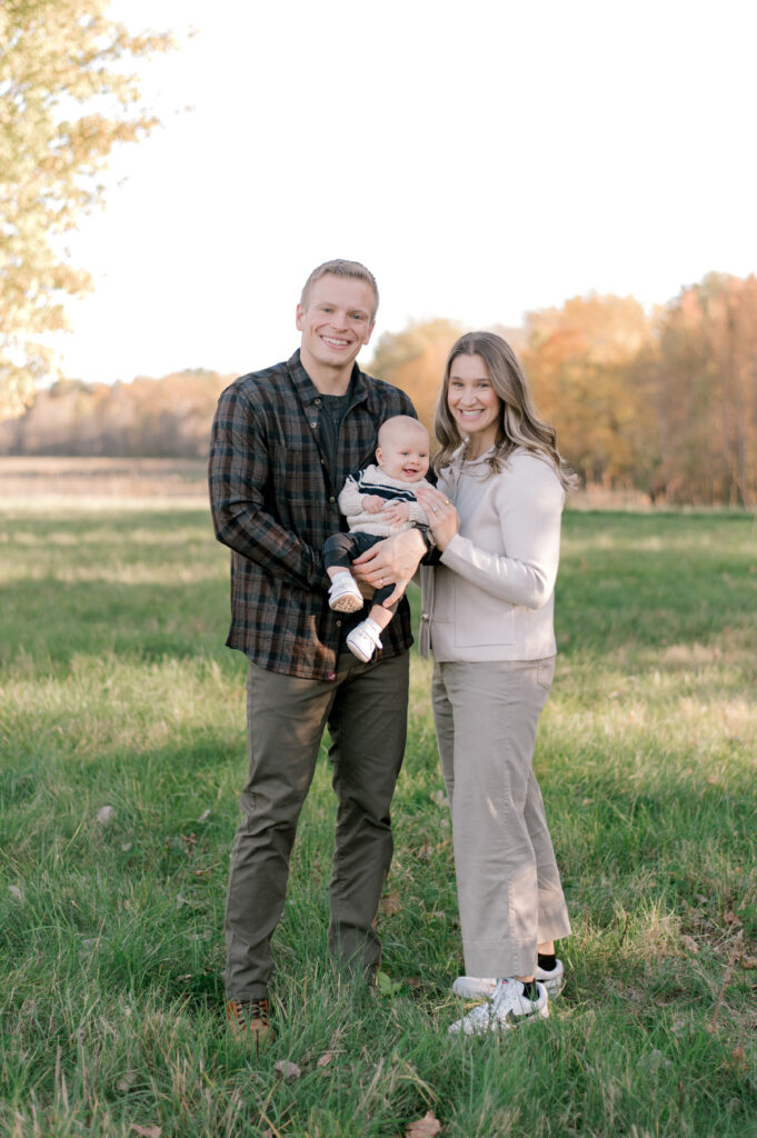 A young family of three, standing in an open field in the beginning of fall, smiling at the camera while mom and dad both hold onto their young, 6 month old son. The family is in a color palette of neautral creams and browns to reflect the season. Photographed by Cleveland Family Photographer, Brittany Serowski Photography.