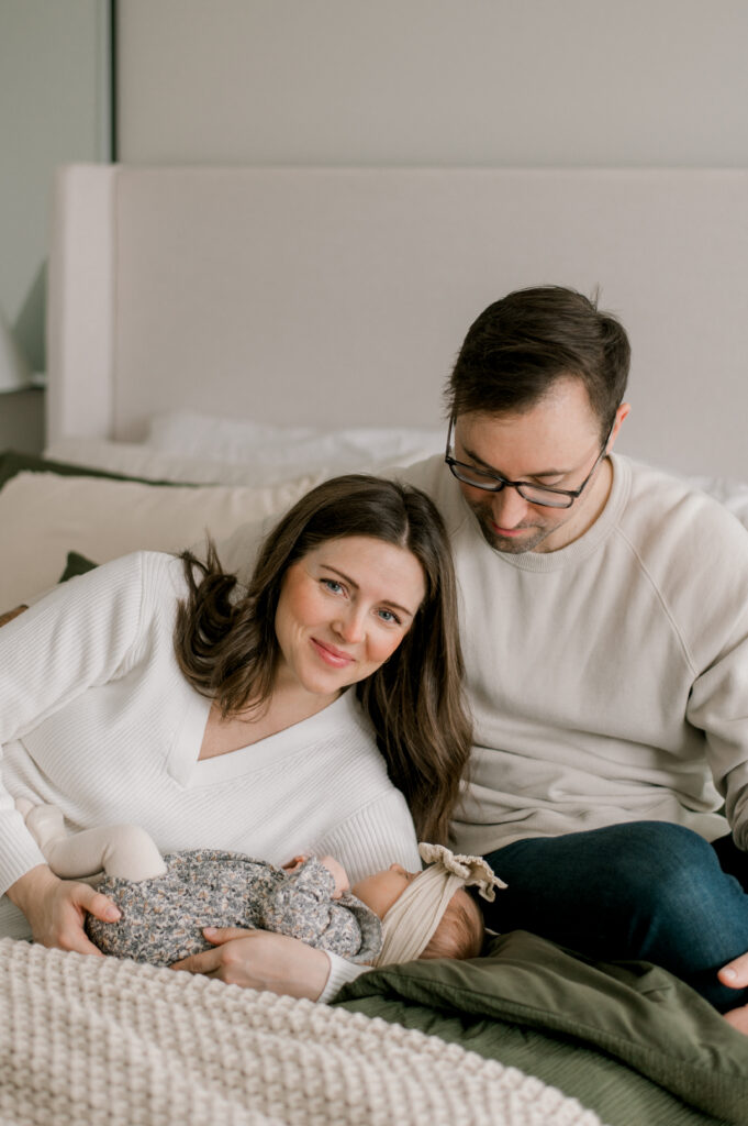 A mom and dad sitting/laying on their bed with their newborn daughter. Their bedding and bedroom colors are neutral in cream as well as their clothing. Mom is smiling and looking at the camera while dad is staring down at his newborn daughter. Photographed by Cleveland Newborn Photographer, Brittany Serowski Photography.