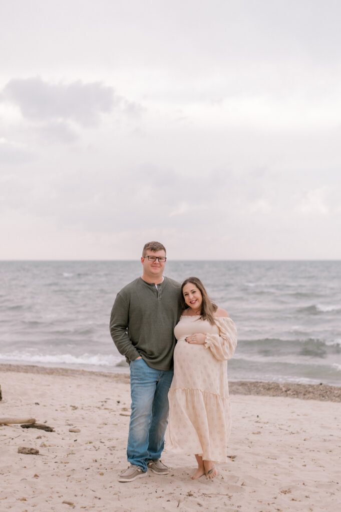 An expecting couple standing side by side, snuggled in together as they smile at the camera ahead. They are standing along the Vermilion beaches in front of Lake Erie. Photographed by Cleveland Maternity Photographer, Brittany Serowski Photography.
