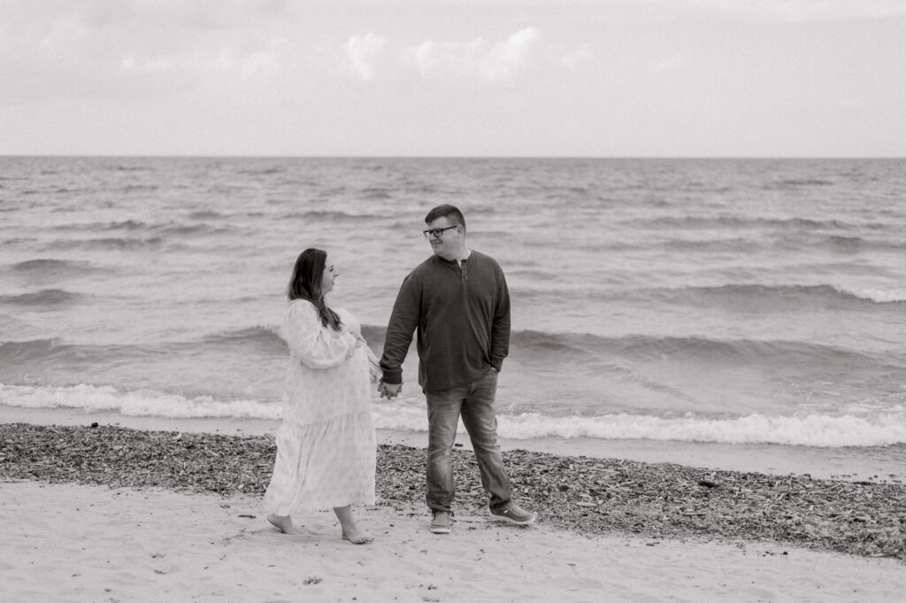 A black and white photo of a couple holding hands who is expecting their first child. They are walking along the shores of Lake Erie in Vermilion, OH. Photographed by Cleveland Maternity Photographer, Brittany Serowski Photography.