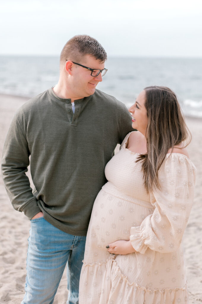 An expecting couple, mom wearing a cream dress and dad in a sage green shirt, smiling at one another as they hold onto the mom's expecting baby bump. They are standing along the shores of Lake Erie in Vermilion, OH. Captured by CLeveland Maternity Photographer, Brittany Serowski Photography.