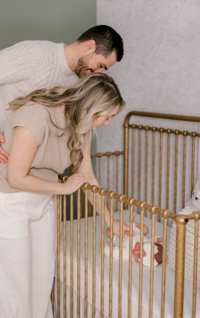 A new mom and dad standing over their newborn daughters crib, holding onto her little hand as they stare lovingly down at her. The crib is a beautiful gold with sage green walls and a cream, floral wallpaper behind. Captured by Cleveland Newborn Photographer, Brittany Serowski Photography.