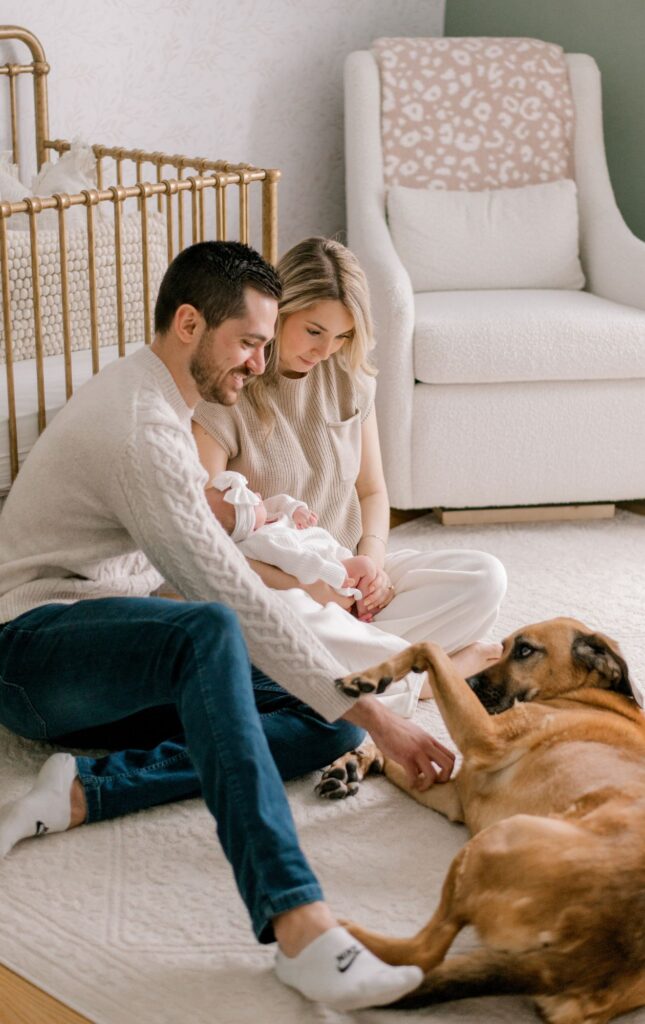 A new mom and dad sitting on the floor of their daughter's nursery while sitting in front of her crib, holding their baby girl and playing with their puppy too. The nursery is decorated in sage green and cream floral wallpaper neutrals. Photographed by Cleveland Newborn Photographer, Brittany Serowski Photography.