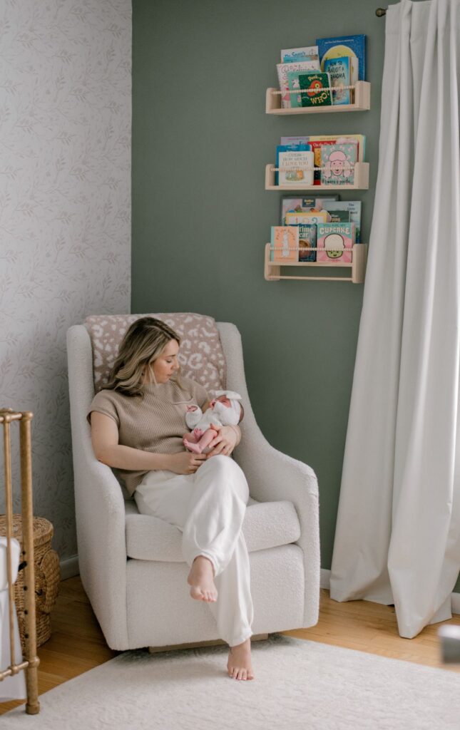 A new mom sitting in a cream boucle rocking chair, holding her newborn daughter. Mom and baby are both in cream colored outfits while they look at one another. The nursery is sage green and cream floral wallpaper. Photographed by Cleveland Newborn Photographer, Brittany Serowski Photography.