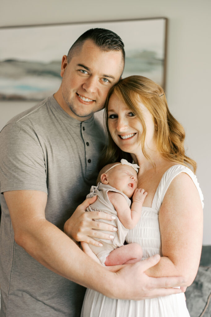 Mother and father holding newborn close, captured by Cleveland newborn photographer, Brittany Serowski Photography. Mother is in a white scrunch dress while dad is in a beige henley shirt. They are smiling and looking at the camera while their newborn daughter sleeps on mom's chest.
